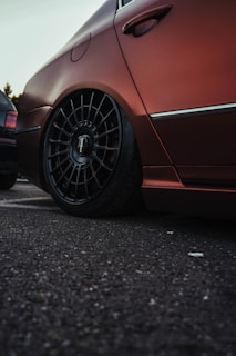 A close-up view of the rear wheel and side panel of a car with a matte finish. The car appears to have aftermarket custom wheels with a multi-spoke design. It is parked on an asphalt surface, and another car with a slightly different color is visible in the background.