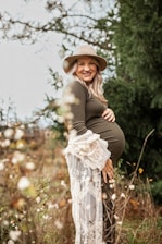 A pregnant woman is smiling and standing in a natural setting surrounded by greenery. She is wearing a hat and a long-sleeved dress with lace details, holding her belly with one hand.