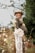 A pregnant woman is smiling and standing in a natural setting surrounded by greenery. She is wearing a hat and a long-sleeved dress with lace details, holding her belly with one hand.