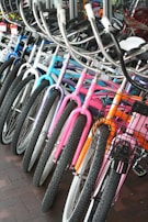 A row of colorful bicycles is lined up, showcasing a variety of designs and hues including blue, purple, pink, and orange. The bicycles are equipped with curved handlebars and some have wire baskets attached to the front. They are positioned on a brick-tiled surface near a glass window.