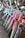A row of colorful bicycles is lined up, showcasing a variety of designs and hues including blue, purple, pink, and orange. The bicycles are equipped with curved handlebars and some have wire baskets attached to the front. They are positioned on a brick-tiled surface near a glass window.