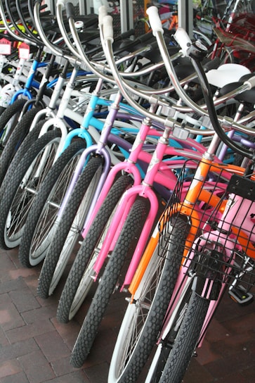 A row of colorful bicycles is lined up, showcasing a variety of designs and hues including blue, purple, pink, and orange. The bicycles are equipped with curved handlebars and some have wire baskets attached to the front. They are positioned on a brick-tiled surface near a glass window.