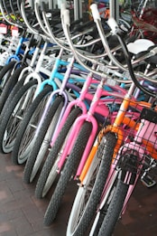 A row of colorful bicycles is lined up, showcasing a variety of designs and hues including blue, purple, pink, and orange. The bicycles are equipped with curved handlebars and some have wire baskets attached to the front. They are positioned on a brick-tiled surface near a glass window.