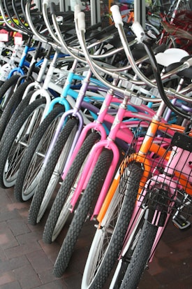 A row of colorful bicycles is lined up, showcasing a variety of designs and hues including blue, purple, pink, and orange. The bicycles are equipped with curved handlebars and some have wire baskets attached to the front. They are positioned on a brick-tiled surface near a glass window.
