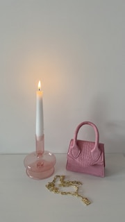 A pink candle holder with a lit white candle sits next to a small pink handbag with crocodile texture on a light surface. A gold chain with a rectangular pendant is placed in front of the handbag.