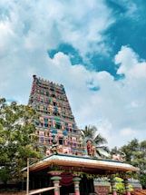 An ornate, colorful Hindu temple tower with intricate carvings and statues rises against a backdrop of a bright blue sky scattered with fluffy white clouds. Lush green trees surround the temple structure.