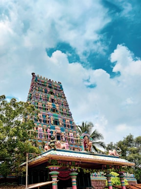 An ornate, colorful Hindu temple tower with intricate carvings and statues rises against a backdrop of a bright blue sky scattered with fluffy white clouds. Lush green trees surround the temple structure.