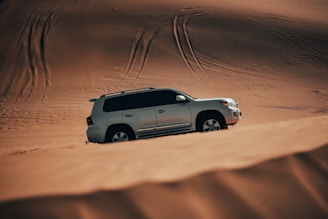 A silver SUV is positioned on a vast expanse of sand dunes. The vehicle appears to be making its way through the undulating terrain, with tire tracks visible in the background. The sand has a smooth texture with varying shades of brown, suggesting a desert environment.