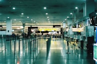 A spacious and brightly lit airport terminal with high ceilings. Multiple service desks line the right side, each equipped with monitors and signage. On the left, rows of seats are visible. The area is reflective with polished tile floors, and overhead lights illuminate the space. A few people are scattered across the terminal, adding a sense of minimal activity.