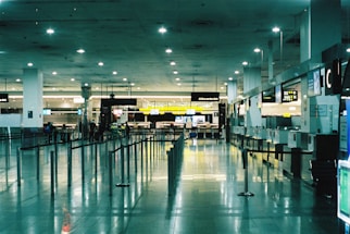 A spacious and brightly lit airport terminal with high ceilings. Multiple service desks line the right side, each equipped with monitors and signage. On the left, rows of seats are visible. The area is reflective with polished tile floors, and overhead lights illuminate the space. A few people are scattered across the terminal, adding a sense of minimal activity.