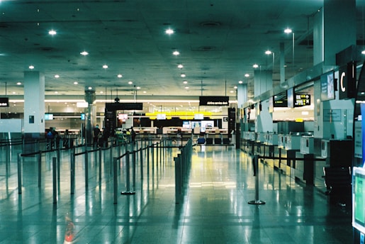 A spacious and brightly lit airport terminal with high ceilings. Multiple service desks line the right side, each equipped with monitors and signage. On the left, rows of seats are visible. The area is reflective with polished tile floors, and overhead lights illuminate the space. A few people are scattered across the terminal, adding a sense of minimal activity.