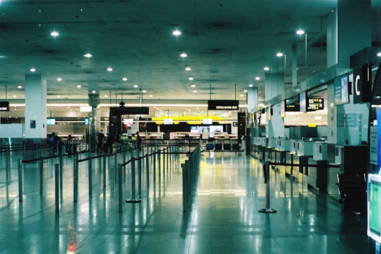 A spacious and brightly lit airport terminal with high ceilings. Multiple service desks line the right side, each equipped with monitors and signage. On the left, rows of seats are visible. The area is reflective with polished tile floors, and overhead lights illuminate the space. A few people are scattered across the terminal, adding a sense of minimal activity.