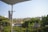 A balcony view features a metal railing and lush greenery in the foreground, with a satellite dish visible. A towel hangs on a clothesline. In the background, there is a landscape with rolling hills and scattered trees under a clear, pale blue sky.