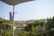 A balcony view features a metal railing and lush greenery in the foreground, with a satellite dish visible. A towel hangs on a clothesline. In the background, there is a landscape with rolling hills and scattered trees under a clear, pale blue sky.