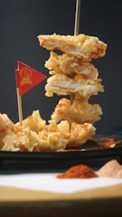 A skewer holds several pieces of deep-fried, crispy food, possibly chicken, with a small red flag in the foreground. The dish is served on a dark plate with a sprinkling of red seasoning. In the background, there is a blurred view of two piles of powdered seasoning, one red and one beige.