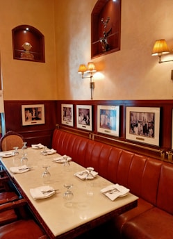 A dining area with a polished marble table set for a meal, complete with glassware and neatly folded napkins. The walls are adorned with framed black and white photographs and decorative wall sconces providing warm lighting. Above the seating area, niches display sculptures.