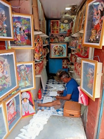 A narrow shop is filled with colorful framed depictions of deities and religious figures. A person is seated on the floor, engaged in creating or crafting, surrounded by art supplies and religious motifs. The space is vibrant and richly decorated with spiritual artwork, suggesting a focus on religious or cultural artifacts.