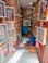 A narrow shop is filled with colorful framed depictions of deities and religious figures. A person is seated on the floor, engaged in creating or crafting, surrounded by art supplies and religious motifs. The space is vibrant and richly decorated with spiritual artwork, suggesting a focus on religious or cultural artifacts.