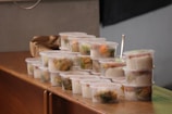 Multiple stackable plastic containers filled with rice and vegetables are arranged on a wooden surface. The containers are transparent, allowing for an easy view of the colorful contents inside. A brown paper bag is seen in the background.