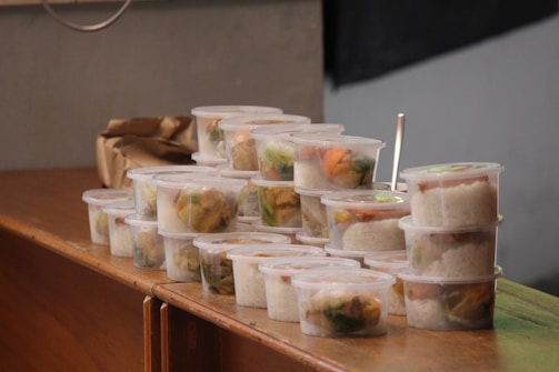 Multiple stackable plastic containers filled with rice and vegetables are arranged on a wooden surface. The containers are transparent, allowing for an easy view of the colorful contents inside. A brown paper bag is seen in the background.