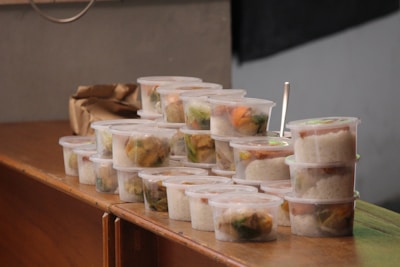 Multiple stackable plastic containers filled with rice and vegetables are arranged on a wooden surface. The containers are transparent, allowing for an easy view of the colorful contents inside. A brown paper bag is seen in the background.
