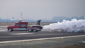 A powerful red truck with checkerboard patterns on its sides races down a runway, emitting a large plume of white smoke from its rear. The truck appears to have jet propulsions, evidenced by the intense smoke and visible jet exhaust. The background is an open, foggy landscape with overcast skies and distant silhouettes of trees and power lines.