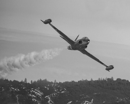 A vintage fighter jet flies at a steep angle, leaving a trail of smoke behind. The aircraft is set against a backdrop of misty clouds, with a forested mountain range below.