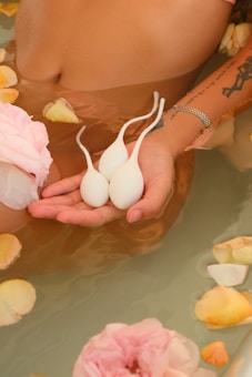 A serene setting in a bath with pink and peach flower petals floating on the water's surface. An individual with tattoos on their arm holds three white teardrop-shaped objects in their hand. The skin has a subtle sheen from the water, contributing to the calm and peaceful atmosphere.