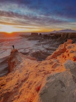 A person stands at the edge of a rocky cliff, overlooking a vast desert landscape. The sky is vibrant with a sunset, casting warm hues over the rugged terrain and creating a dramatic contrast with the blue sky. Layers of clouds add texture to the scene, while distant mountains are visible on the horizon.