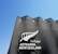 A modern architectural structure with a triangular roof design features the text 'Aotearoa New Zealand' accompanied by a stylized silver fern symbol. The building facade is dark with a textured pattern and contrasts against a clear blue sky.