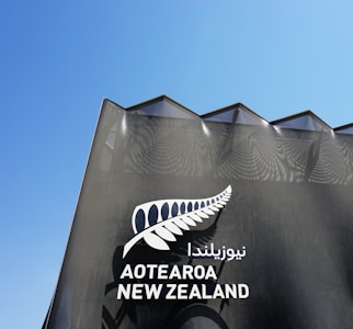 A modern architectural structure with a triangular roof design features the text 'Aotearoa New Zealand' accompanied by a stylized silver fern symbol. The building facade is dark with a textured pattern and contrasts against a clear blue sky.