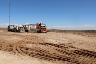 A barren landscape with two large trucks hauling heavy machinery across a dusty expanse. The ground is marked with deep tire tracks, and there is a tall, thin tower in the background against a clear blue sky.