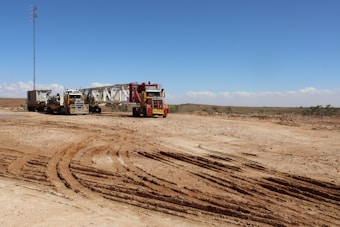 A barren landscape with two large trucks hauling heavy machinery across a dusty expanse. The ground is marked with deep tire tracks, and there is a tall, thin tower in the background against a clear blue sky.
