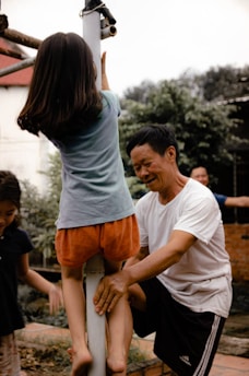 A girl is climbing a pole with the assistance of an older man who is supporting her from behind. They are outdoors, possibly in a backyard, with another child and a brick wall in the background. The scene suggests a playful and familial atmosphere.