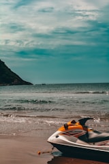A jet ski is parked on the sandy shore of a beach with calm waves in the background. The ocean extends to the horizon, meeting a partly cloudy sky. There is a distant view of a green-covered hill on the left side, and a small boat can be seen far out in the water.