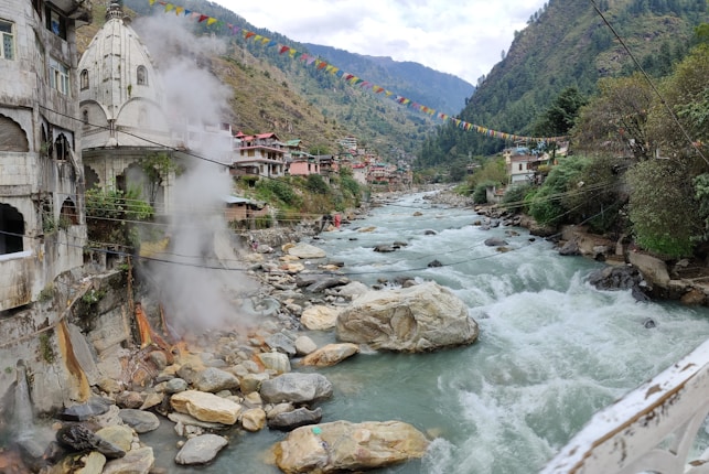 A scenic mountain river flows through a valley bordered by buildings and lush greenery. Steam rises near a stone structure with an ornate temple-like design. Colorful prayer flags are strung across the river, adding a cultural touch to the landscape. The river is lined with large rocks and boulders, creating a dynamic natural scene.