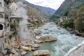 A scenic mountain river flows through a valley bordered by buildings and lush greenery. Steam rises near a stone structure with an ornate temple-like design. Colorful prayer flags are strung across the river, adding a cultural touch to the landscape. The river is lined with large rocks and boulders, creating a dynamic natural scene.