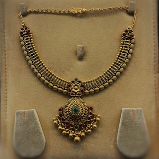 An ornate gold necklace with intricate detailing and embedded gemstones is displayed against a beige background. The necklace features a series of bead-like structures forming the main chain, while a large central pendant is adorned with a combination of green and red gems, accentuated with floral motifs.