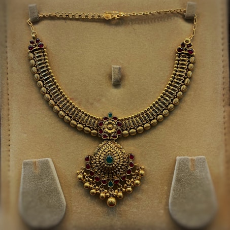 An ornate gold necklace with intricate detailing and embedded gemstones is displayed against a beige background. The necklace features a series of bead-like structures forming the main chain, while a large central pendant is adorned with a combination of green and red gems, accentuated with floral motifs.