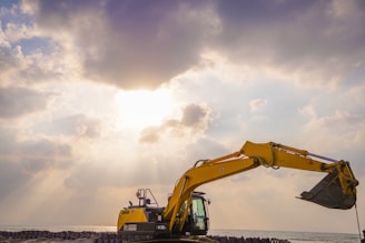 A yellow excavator with an extended arm and bucket stands against a dramatic sky with sun rays peeking through dense clouds. Below, there are concrete barriers lined up along the shoreline, with the sea visible in the background.