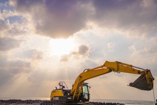 A yellow excavator with an extended arm and bucket stands against a dramatic sky with sun rays peeking through dense clouds. Below, there are concrete barriers lined up along the shoreline, with the sea visible in the background.