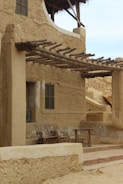 An adobe-style building with rustic wooden beams extending as a pergola. A set of wicker chairs and a table are placed under the pergola on a small porch, surrounded by earthen walls with texture. A window with green shutters is visible.