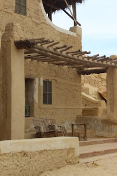 An adobe-style building with rustic wooden beams extending as a pergola. A set of wicker chairs and a table are placed under the pergola on a small porch, surrounded by earthen walls with texture. A window with green shutters is visible.
