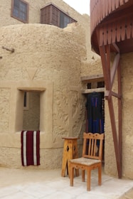 A traditional structure made of mud bricks with wooden elements. A carved wall is featured, and a window with a red and white cloth draped over the ledge. In front of the building, there is a wooden chair and a small table. Hanging next to the door is a dark woven garment with blue and red patterns.