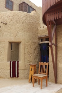 A traditional structure made of mud bricks with wooden elements. A carved wall is featured, and a window with a red and white cloth draped over the ledge. In front of the building, there is a wooden chair and a small table. Hanging next to the door is a dark woven garment with blue and red patterns.