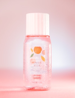 A clear plastic bottle contains a pinkish liquid and has a white cap. The label features an illustration of peaches and text in both Chinese and English, indicating it is a Peach Oolong Mouthwash. The bottle is adorned with droplets, giving a fresh, cool look while sitting against a pink gradient background.