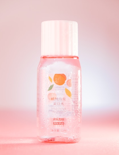 A clear plastic bottle contains a pinkish liquid and has a white cap. The label features an illustration of peaches and text in both Chinese and English, indicating it is a Peach Oolong Mouthwash. The bottle is adorned with droplets, giving a fresh, cool look while sitting against a pink gradient background.