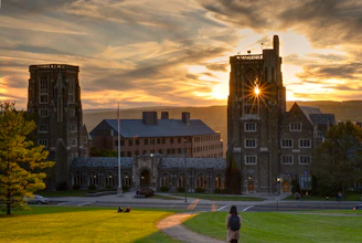 A historic university building with large, stone towers is set against a dramatic sunset sky. The sunlight gleams through the upper part of one tower, creating a picturesque effect. In the foreground, a person with a backpack stands on a grassy hill, looking towards the building. A few people are sitting on the grass, and an American flag is visible.