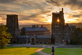 A historic university building with large, stone towers is set against a dramatic sunset sky. The sunlight gleams through the upper part of one tower, creating a picturesque effect. In the foreground, a person with a backpack stands on a grassy hill, looking towards the building. A few people are sitting on the grass, and an American flag is visible.