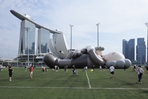 A large inflatable figure resembling a cartoon character lies on a grassy field. People are walking around it, taking photos and observing. In the background, there are tall, modern skyscrapers, including a distinctive building with a curved structure on top.
